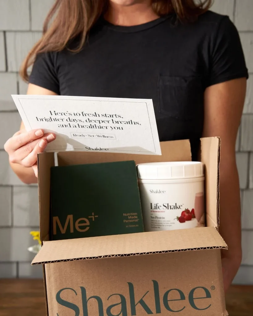 Person holding a cardboard Shaklee box with wellness items, including a Life Shake canister and a green Me+ booklet, plus a card with a health message.